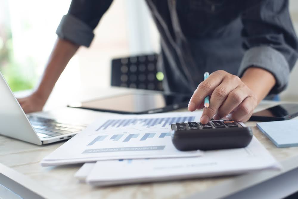 Business owner standing at a desk, reviewing papers, and using a calculator for financial management.